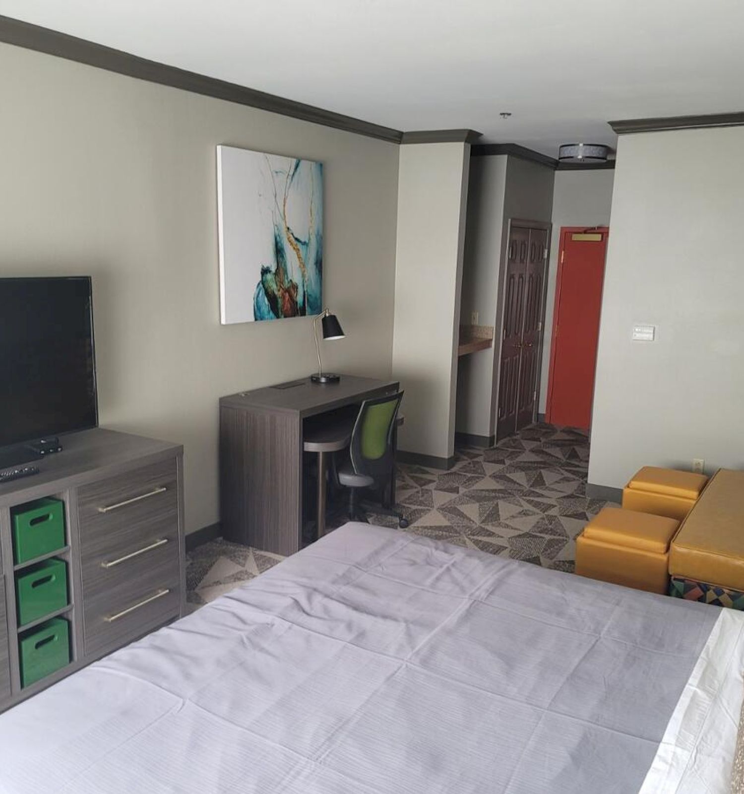 A modern hotel room with a bed, TV, dresser, desk, chair, yellow ottomans, abstract art, and patterned carpet.