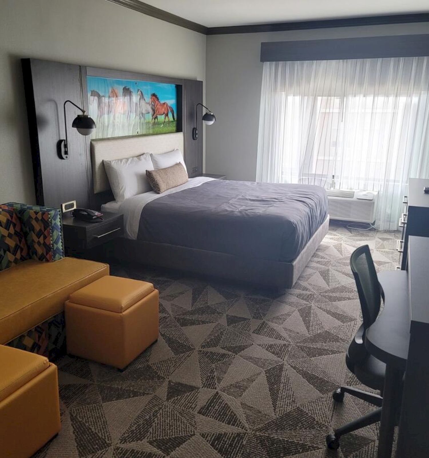 A modern hotel room featuring a bed, a side table, a couch, two ottomans, a desk with a chair, and a window with sheer curtains.