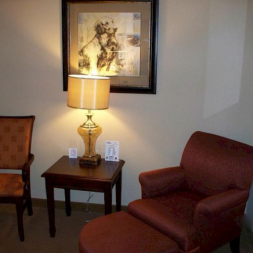 The image shows a cozy room with a red armchair, a matching ottoman, a wooden side table with a lamp, and a framed picture on the wall.