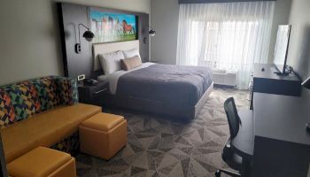 A modern hotel room with a bed, sofa, two ottomans, desk, chair, and TV. Large window with curtains, carpeted floor, and wall art above the bed.