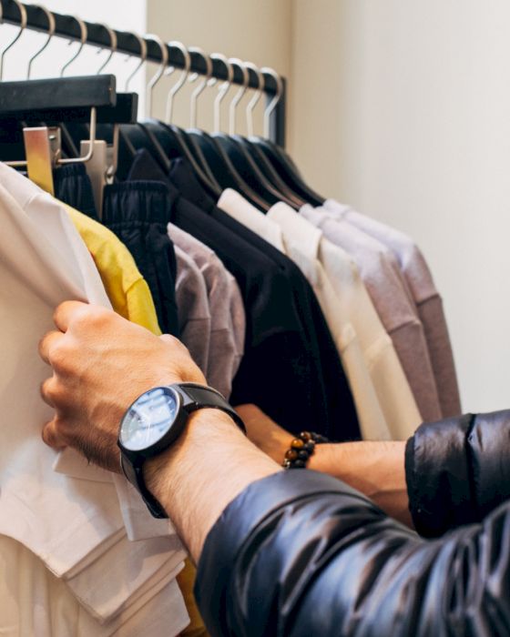 A person wearing a watch browses through a rack of clothes, holding a white t-shirt with "NO FUN" printed on it, in a clothing store.